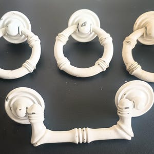 Puede incluir: Un juego de cuatro tiradores de mueble en color blanco roto con una textura inspirada en el bambú. Tres piezas son tiradores de anilla circulares con placas de montaje redondas y una pieza es un tirador horizontal con dos placas.