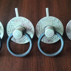 Puede incluir: Cuatro tiradores de cajón antiguos de latón con anillos de hierro gris oscuro. Cada tirador tiene una placa trasera circular con un diseño de abanico y floral, un centro cilíndrico y un pequeño vástago vertical. Los tiradores están dispuestos sobre una superficie de madera oscura.