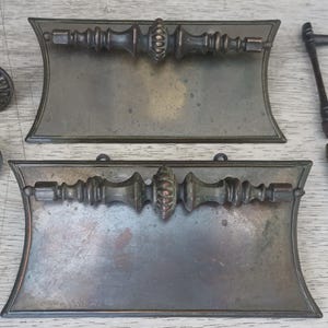 May include: Two rectangular bronze drawer pulls with ornate central spindles, two circular bronze knobs with radial patterns, and two slender metal handles with attached mounting screws, all featuring a dark, aged metallic finish.