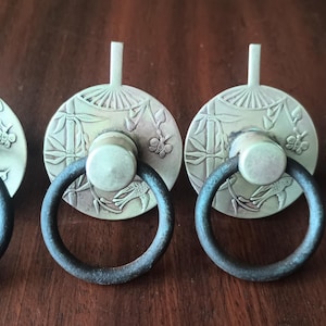 May include: Four antique bronze cabinet pulls with a circular design featuring bamboo and floral patterns. Each pull has a dark ring handle and a small, vertical post. The pulls are arranged in a row on a dark wooden surface.