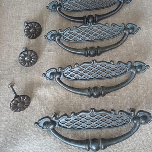 May include: Four dark metal drawer pulls with a lattice-patterned backplate and a curved handle, accompanied by three matching circular knobs with a floral petal design. The hardware features a weathered finish with visible dark gray tones.