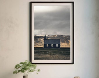 Pode incluir: Fotografia emoldurada de uma paisagem com uma pequena igreja escura com um campanário, em um fundo de colinas e um céu nublado. A imagem é exibida acima de um armário de madeira com um vaso de vegetação e um objeto decorativo.