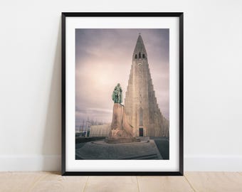 Pode incluir: Uma fotografia a preto e branco de uma igreja moderna e alta com uma estátua à sua frente. A igreja tem um telhado pontiagudo e um relógio na fachada. A estátua é de um homem em pé sobre um pedestal.