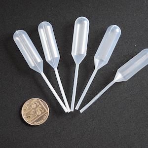 May include: Five clear plastic droppers with a bulbous tip and a long, thin stem. The droppers are arranged in a fan shape on a black surface. A silver coin is in the lower left corner of the image.