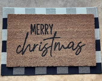 May include: A rectangular doormat with the words "MERRY christmas" in black script. The doormat is brown and sits on a gray and black checkered rug. The rug has a black border.