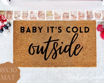 May include: A rectangular doormat with the text "BABY IT'S COLD outside" in black. The mat is 18 x 30 inches and made of a natural fiber material. Festive decorations, including pinecones and ornaments, are visible around the mat.