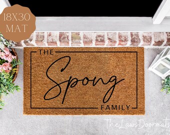May include: A rectangular doormat with the text "The Spong Family" in black script. The mat is 18x30 inches and made of a natural brown material. The text is enclosed in a black rectangular border.