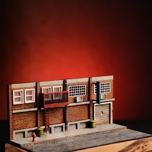 May include: A miniature model of a brick building with a red fire escape, a balcony, and a garage door. The model is set on a wooden base with a gray cobblestone street.