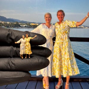 May include: Miniature figurines of two women in dresses, held in a gloved hand, are positioned in front of two women wearing similar dresses. The background shows a coastal scene with a pier and ocean. The dresses feature floral patterns.