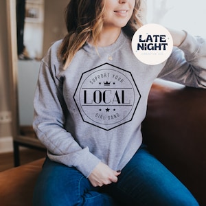 Può includere: Una felpa grigia con la scritta "SUPPORT YOUR LOCAL GIRL GANG" in un design ottagonale nero. La felpa presenta anche il logo Late Night Creative Co. in un cerchio bianco.