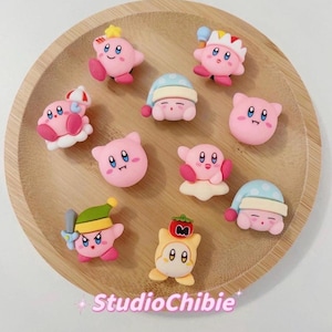 May include: A collection of pink Kirby character figurines with various accessories, including hats, a sword, and a cloud. The figurines are arranged on a round wooden tray. The text "StudioChibie" is at the bottom.