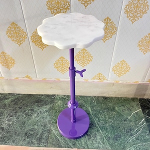 May include: A small, decorative table with a white, scalloped marble top. The table has a vibrant purple, adjustable metal stem and base. The base is round. The table is designed for display or decorative purposes.
