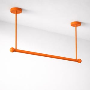 Puede incluir: Perchero naranja montado en el techo. El perchero presenta una barra horizontal suspendida del techo por dos varillas verticales y soportes circulares. Los extremos de la barra horizontal tienen pequeños detalles esféricos. El perchero está pintado de color naranja liso.