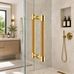 Puede incluir: Un tirador de puerta de ducha dorado con un agarre texturizado. El tirador es cilíndrico con un patrón moleteado y está fijado a la puerta de cristal con herrajes dorados. El baño tiene una pared de baldosas de mármol y accesorios dorados.