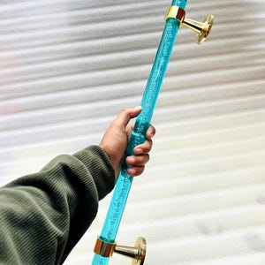 May include: A clear, turquoise door handle with gold-coloured accents. The handle is cylindrical with a bubble-like texture and is mounted on two gold-coloured circular bases. The handle is held in a hand.