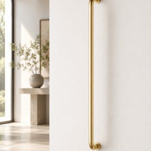 May include: A gold-coloured, vertical door pull or handle fixed to a white wall. The handle features a curved design at each end and a straight, cylindrical central section. The text "ModernHomecor" is at the bottom of the image.