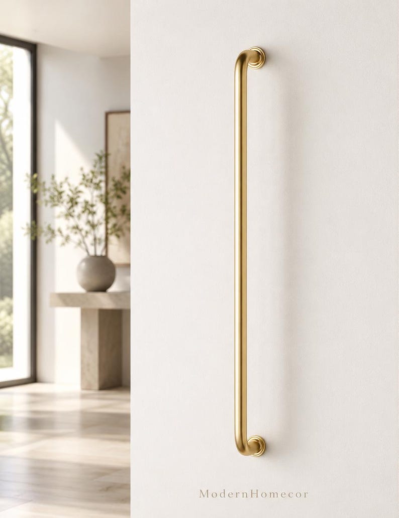May include: A gold-coloured, vertical door pull or handle fixed to a white wall. The handle features a curved design at each end and a straight, cylindrical central section. The text "ModernHomecor" is at the bottom of the image.