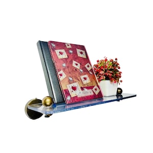 May include: A clear glass shelf with brass accents holds a red and gold journal and a small potted plant with red flowers. The shelf is mounted to a white wall.