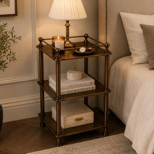 May include: A bronze-coloured three-tiered side table with a lamp, books, and decorative items. The lamp has a white shade and sits on the top shelf, along with a candle and a small dish. The second shelf holds books, and the bottom shelf has a box.