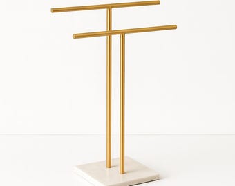 Può includere: Un espositore per gioielli in metallo color oro. Presenta due barre orizzontali per appendere i gioielli, sostenute da montanti verticali. La base è in marmo bianco sporco quadrato. Per collane, bracciali e altri accessori.