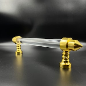 May include: A gold-toned door handle with a clear acrylic center bar. The handle has a pointed end and a decorative, fluted base.
