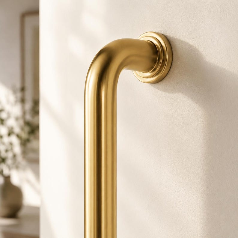 May include: A gold-coloured, curved grab bar fixed to a white wall. The bar features a circular mounting plate and a smooth, cylindrical design. The lighting accentuates the metallic finish, suggesting a modern bathroom fitting.