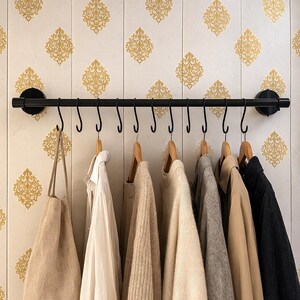 May include: A black metal wall-mounted coat rack with multiple hooks, holding various garments on wooden hangers. The items include a beige tote bag, a white shirt, sweaters, and a dark jacket. The wall behind the rack has a cream and gold damask pattern.