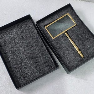 May include: A rectangular magnifying glass with a gold-colored frame and handle rests in a black box with a foam insert. The magnifying glass is designed for reading and detailed viewing. The box is open, revealing the magnifying glass.