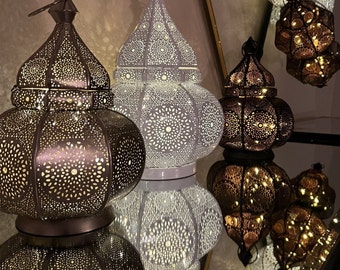 May include: Ornate metal lanterns in bronze and white, featuring detailed cut-out designs. The lanterns are illuminated internally, producing a warm light. The lanterns are displayed on a reflective surface, with more lanterns suspended in the background.