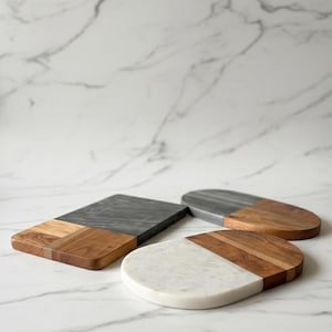 May include: Three serving boards with a two-tone design. One is rectangular with a gray marble top and wood base. The other two are oval-shaped, one with a gray marble top and wood base, and the other with a white marble top and wood base.
