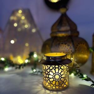 May include: A decorative lantern with a geometric pattern, casting a warm glow. The lantern is gold-colored with a dark brown handle and top. Other lanterns and string lights are in the background, creating a soft, ambient light.