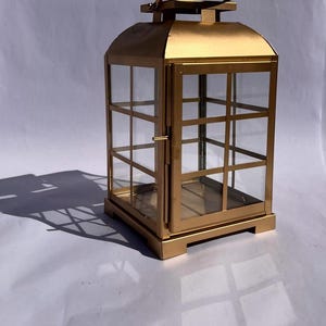 May include: A gold-colored metal lantern with clear glass panels. The lantern has a rectangular base and a handle on top. The glass panels are divided into a grid pattern. The lantern is casting a shadow on a white surface.