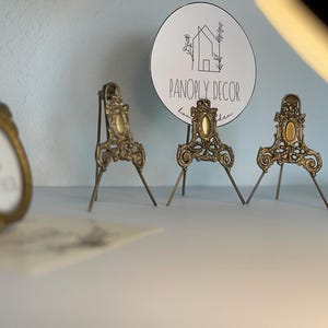 May include: Three ornate, antique-style brass metal easels with intricate scrollwork and a central oval accent. One easel holds a circular white sign featuring a line-art house illustration and the text PANOPLY DECOR in black lettering.