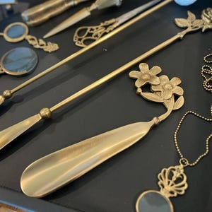 May include: A collection of brass-toned metal accessories including two long-handled shoehorns, one topped with a rose and the other with two flowers, alongside magnifying glasses with ornate handles and a pendant on a beaded chain.