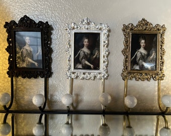 Op de afbeelding: Drie sierlijke fotolijsten met haken aan de onderkant. De lijsten zijn zwart, zilver en goud. Elke lijst bevat een portret van een vrouw in een witte jurk.