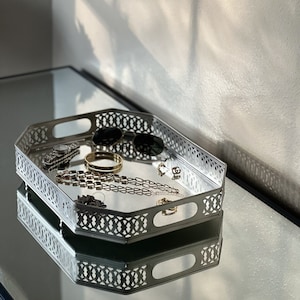 Puede incluir: Una bandeja de joyería octogonal plateada con un diseño calado decorativo. La bandeja contiene varios accesorios, incluyendo gafas de sol, un reloj, pulseras, collares y pendientes. La bandeja está sobre una superficie reflectante.