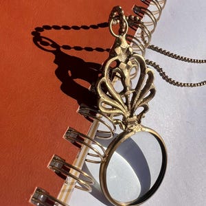 May include: A vintage-style brass magnifying glass pendant with a decorative floral design. The pendant hangs on a delicate chain and rests on a spiral-bound notebook. The notebook is open to a page with the word "QUALITY" printed on it.