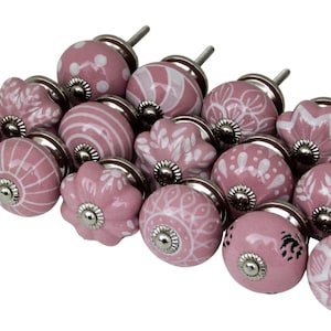 Puede incluir: Un conjunto de pomos de cerámica rosa con diversos diseños blancos. Los pomos, de unos 3 cm de diámetro, presentan patrones de rayas, puntos y florales. Cuentan con una base y un vástago de metal plateado.