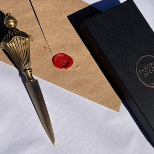 May include: A gold-colored letter opener with a decorative handle, next to a brown envelope sealed with red wax. A black box with the text "PANOPLY DECOR HOME & GARDEN" is also present.