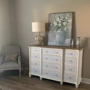 May include: A white dresser with multiple drawers, a wooden top, and a decorative lamp. A framed floral painting and a striped armchair are also visible. The room has a neutral color palette.