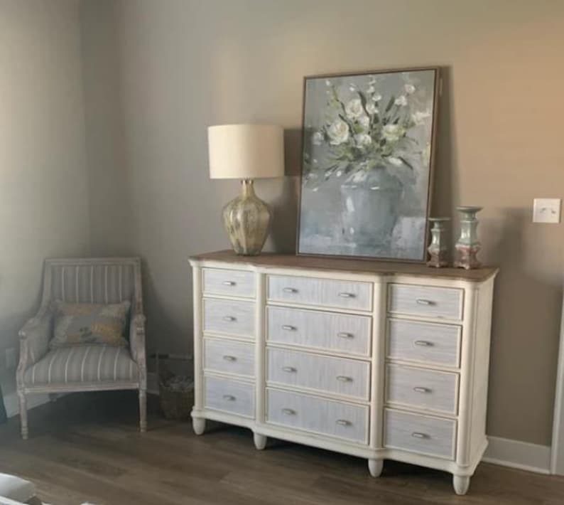 May include: A white dresser with multiple drawers, a wooden top, and a decorative lamp. A framed floral painting and a striped armchair are also visible. The room has a neutral color palette.