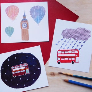 Può includere: Quattro illustrazioni ad acquerello su cartoncino bianco. La prima illustrazione mostra un autobus a due piani rosso che guida attraverso un cielo notturno stellato. La seconda illustrazione mostra un autobus a due piani rosso che guida attraverso una giornata piovosa. La terza illustrazione mostra una mongolfiera blu e una torre dell'orologio in mattoni rossi. La quarta illustrazione mostra una mongolfiera rosa.