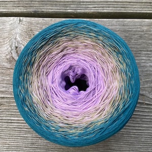 May include: A round cake of yarn featuring a gradient color transition. The center is a light lavender, which shifts outward through a soft beige and tan transition zone, finally reaching a deep, saturated teal blue at the outer edge.