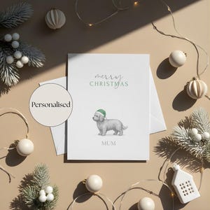 Puede incluir: Una tarjeta de Navidad blanca con las palabras "merry CHRISTMAS" en verde. La tarjeta presenta una ilustración en acuarela de un perro con un sombrero verde. La palabra "MUM" está impresa debajo.
