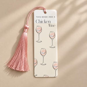 May include: A rectangular bookmark with a cream background featuring a repeating pattern of line-art wine glasses with pink accents. The text reads THIS BOOK AND A Chicken WINE. A dusty pink silk tassel is attached to the top with a matching cord.