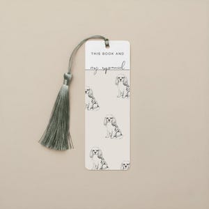 May include: A rectangular bookmark with a light beige background features a repeating pattern of Cavalier King Charles Spaniel illustrations. The text "THIS BOOK AND my spaniel" is printed at the top. A sage green tassel hangs from the top.