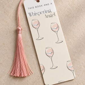 May include: A rectangular bookmark with a cream-coloured background featuring a repeating pattern of line-art wine glasses with pink accents. The text reads THIS BOOK AND A Whispering Angel. A pink silk tassel is attached to the top with a cord.