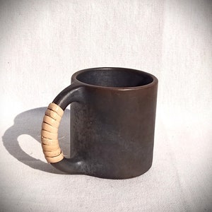 Puede incluir: Una taza de cerámica marrón oscuro con un asa tejida de color beige. La taza tiene un aspecto rústico y hecho a mano.