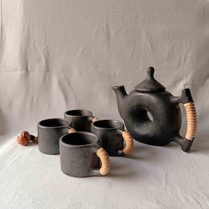 Op de afbeelding: Een zwarte theepot van keramiek met een uniek donutvormig ontwerp en een geweven handvat, samen met vier bijpassende kopjes met geweven handvatten.