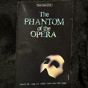 May include: A black theater program for The Phantom of the Opera, featuring the title in white and blue lettering, a white mask graphic, and the text Theater League of Utah, March 28 - July 13, 1996, Salt Lake City, Utah printed on the front cover.